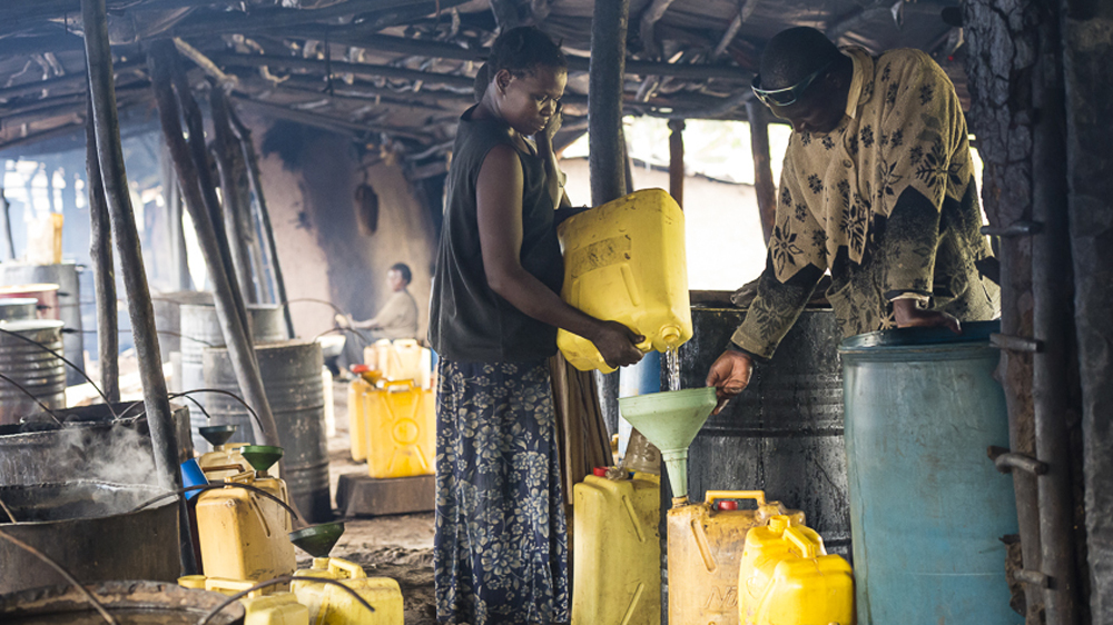 One of Imelda's employees fills up a customer's jerry can with a freshly made batch of waragi [AJ Heath/Al Jazeera]
