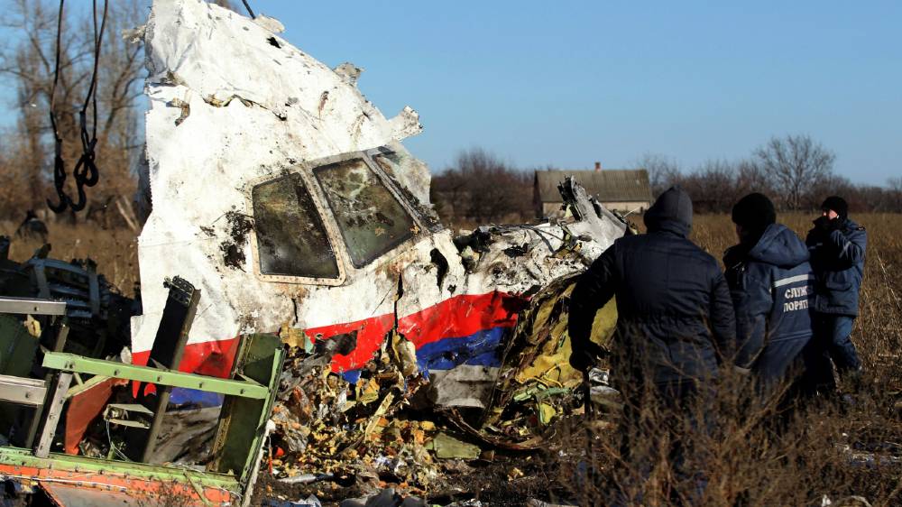 Two workers coming up to front of plane wreck (white with red and blue stripe at bottom) surrounded by debris