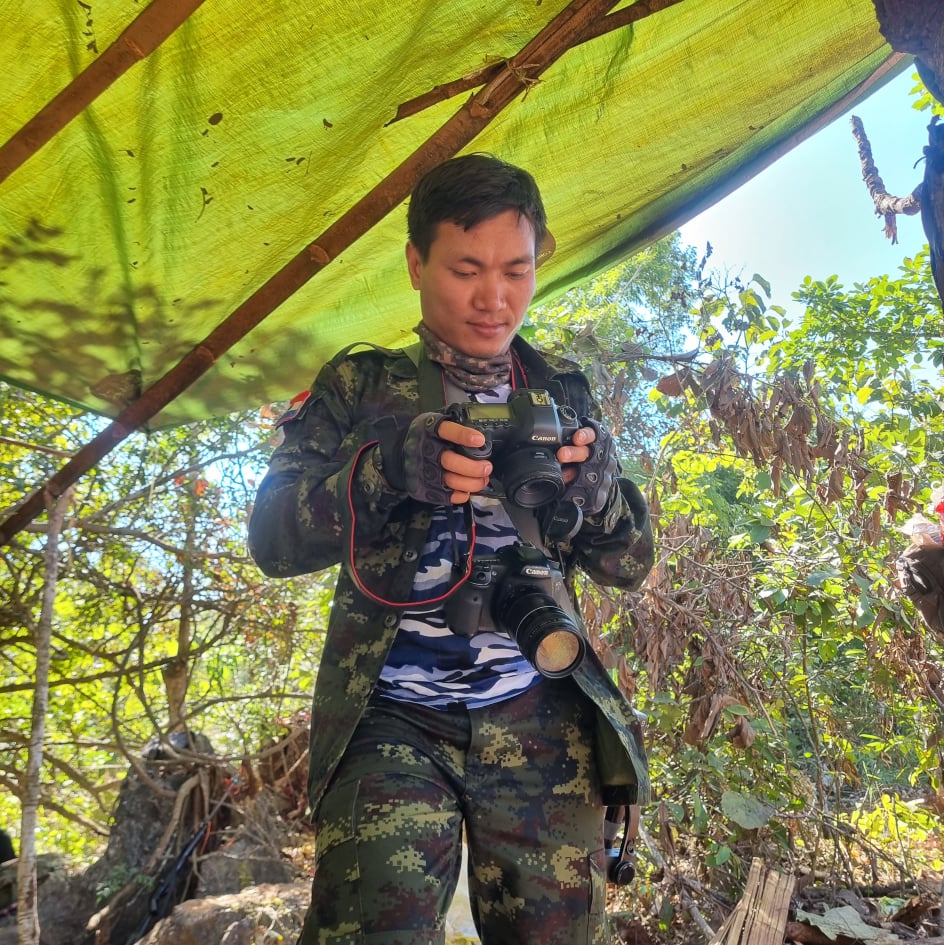 A portrait of Khun Nan Nan standing under a green canopy in the forest, examining his camera.