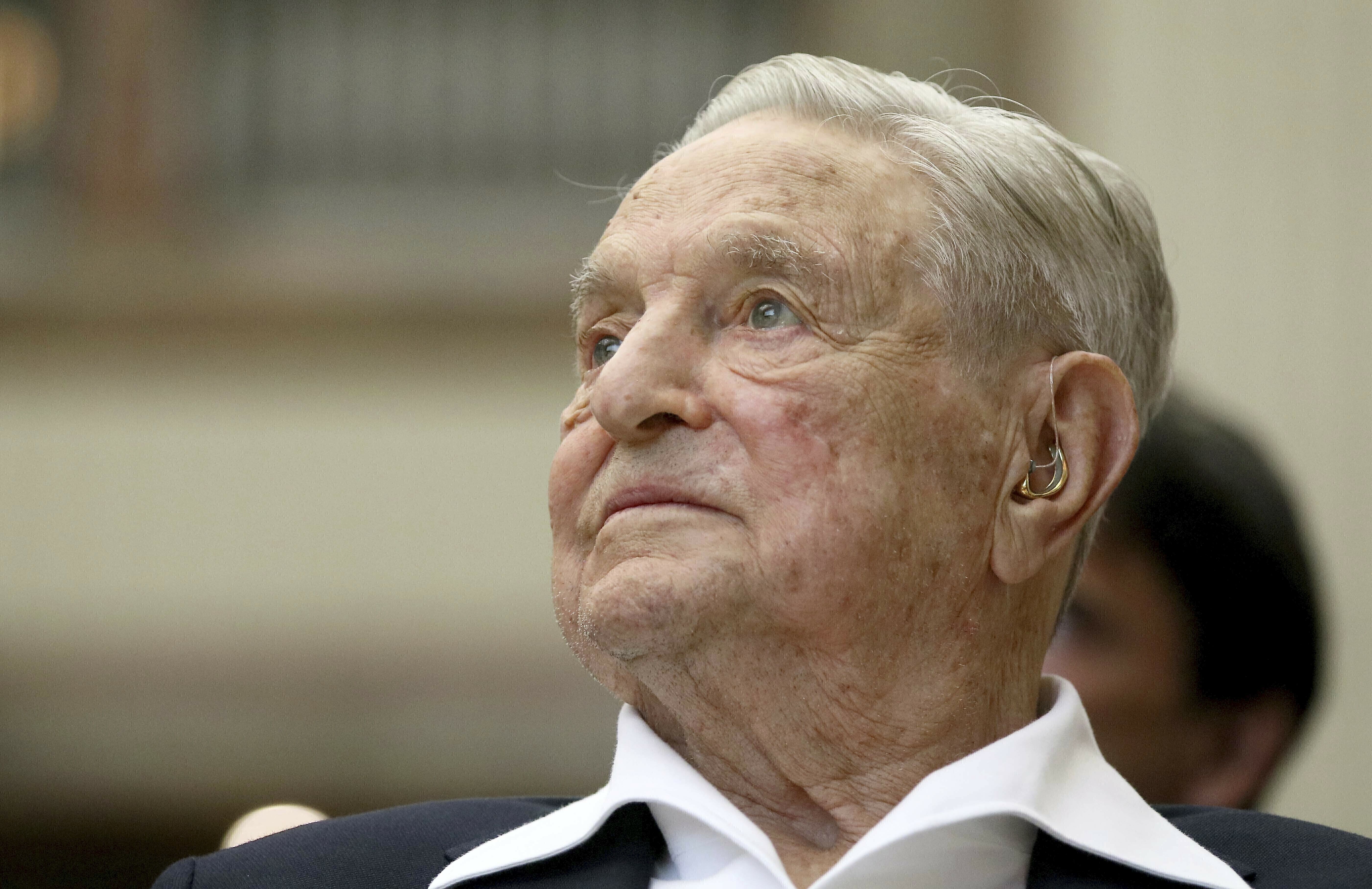 A portrait of George Soros. He is wearing a white open-necked shirt and black jacket. He is listening to someone speaking.
