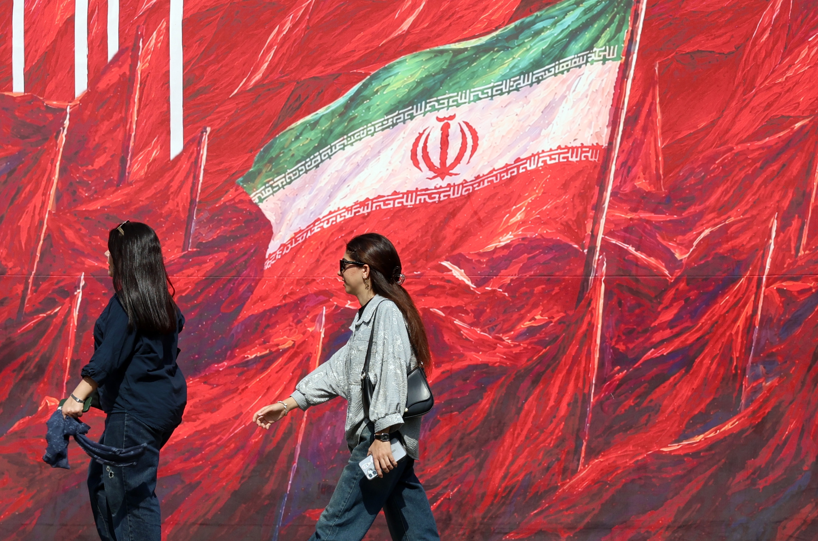 Iranian women walk past a banner featuring Irans national flag in a street in Tehran